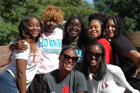 A group of seven women are smiling together outdoors in casual clothing, enjoying a sunny day.